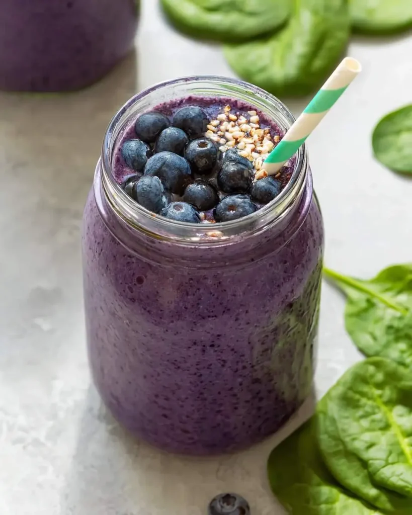A mason jar filled with purple blueberry spinach smoothie topped with fresh blueberries, hemp seeds, and a striped straw.