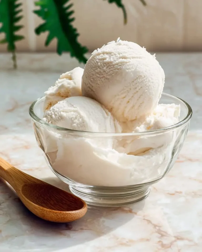 Scoops of creamy coconut milk ice cream served in a clear glass bowl with a wooden spoon beside it.