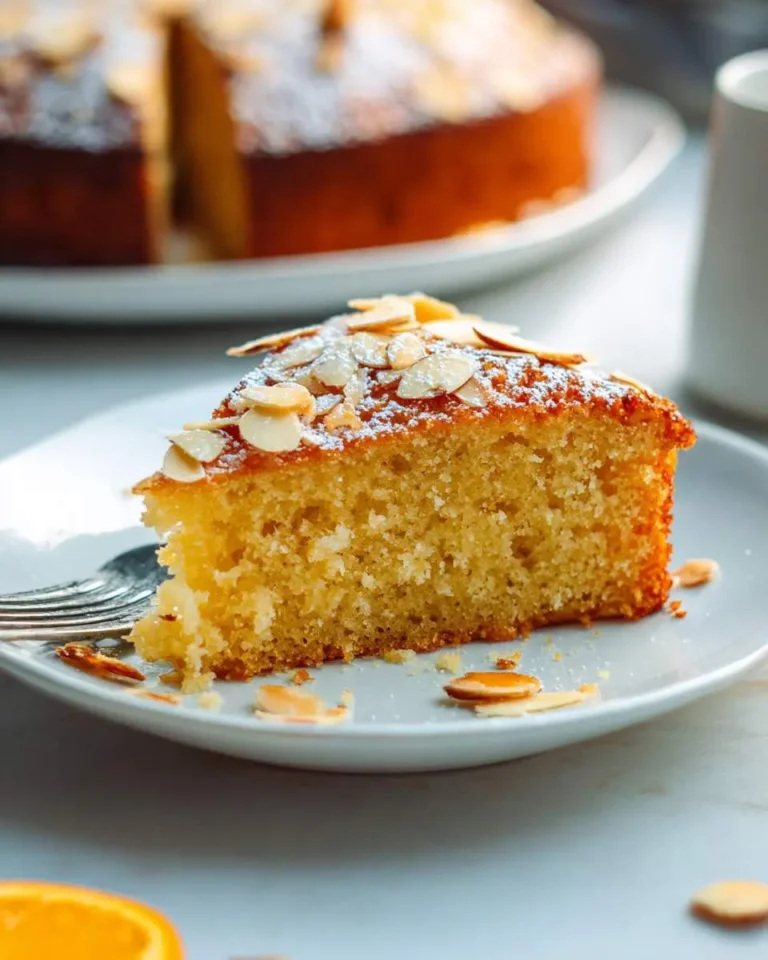 A slice of orange almond cake topped with sliced almonds and powdered sugar on a white plate with a fork.