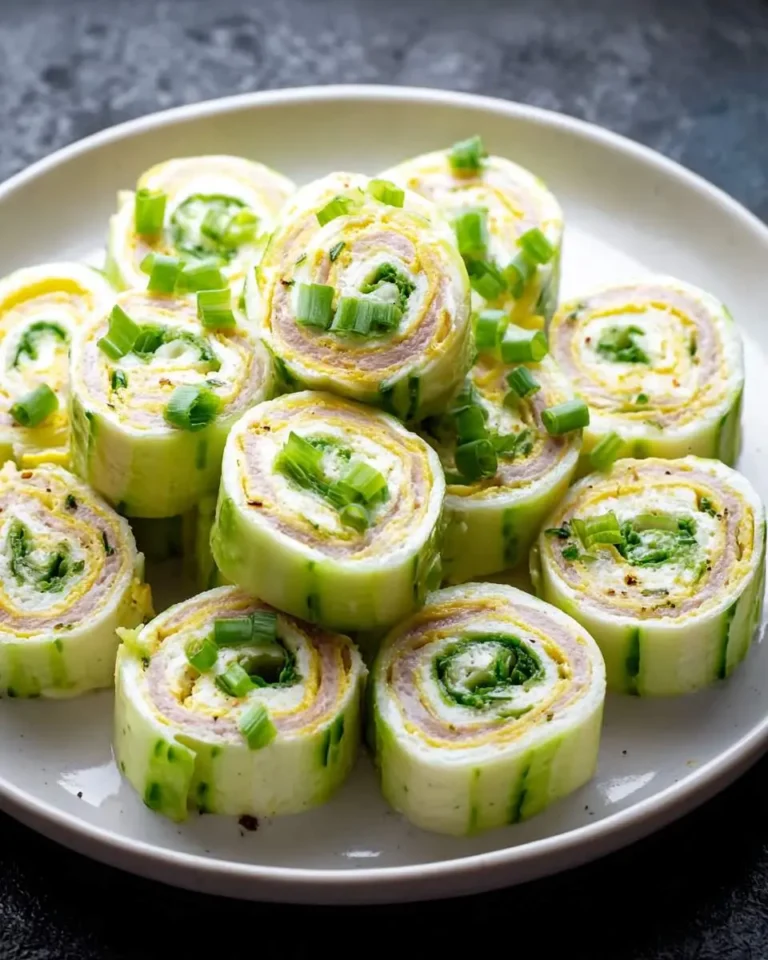 A white plate stacked with Pickle Pinwheels, featuring rolled pickles with cream cheese, meat, and green onion garnish.