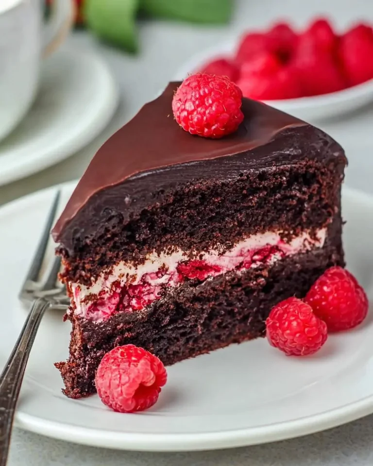 A slice of raspberry chocolate cake with rich ganache topping and fresh raspberries on a white plate.