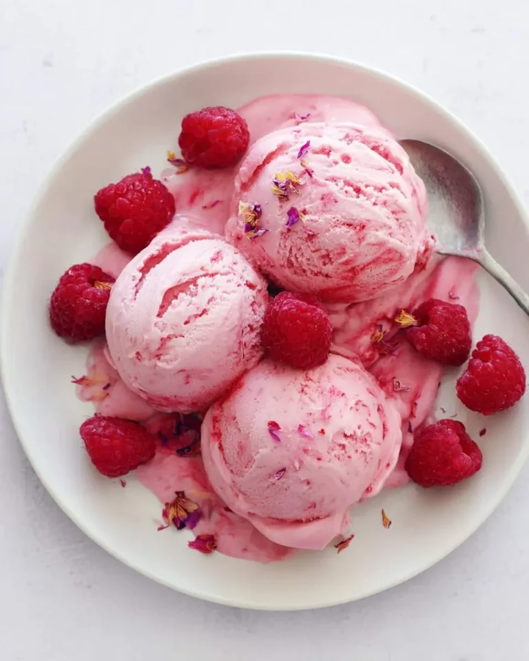 Three scoops of pink raspberry rosewater ice cream in a white bowl with fresh raspberries and edible flower petals.