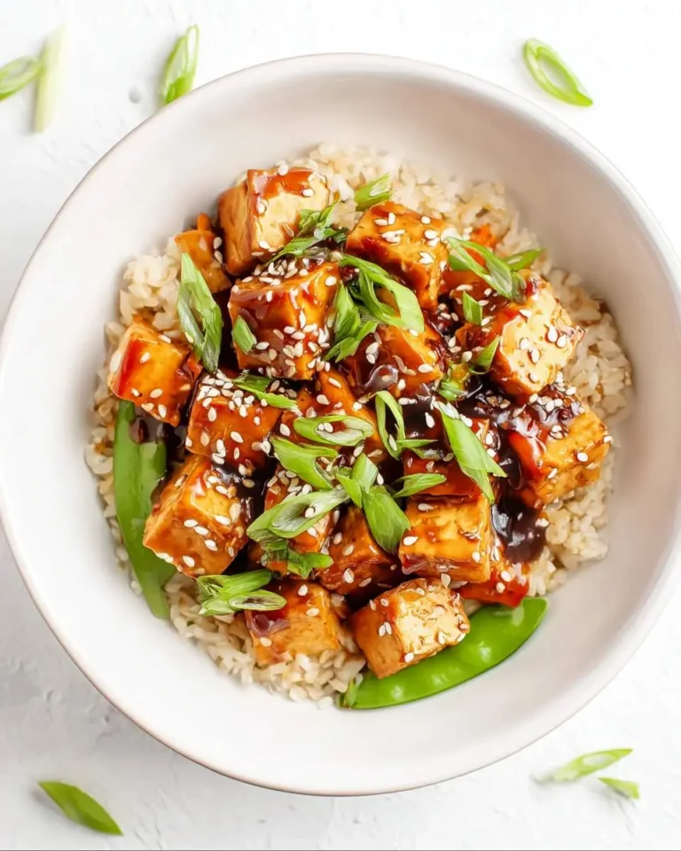 A sesame ginger tofu bowl with crispy tofu cubes glazed in sauce, served over brown rice and topped with green onions and sesame seeds.