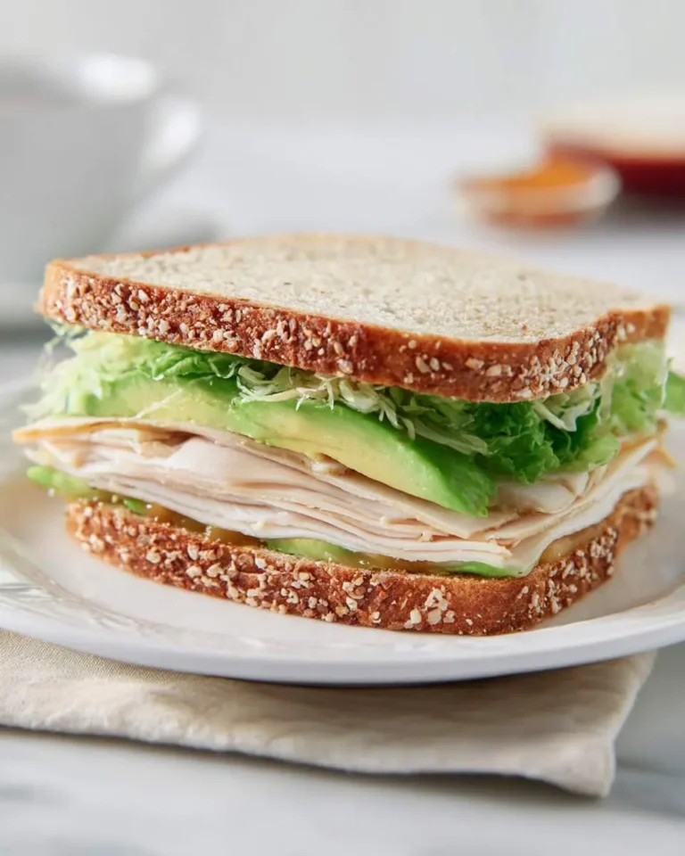 A fresh Turkey Avocado Sandwich with sliced turkey, avocado, lettuce, and sprouts on whole grain bread served on a white plate.
