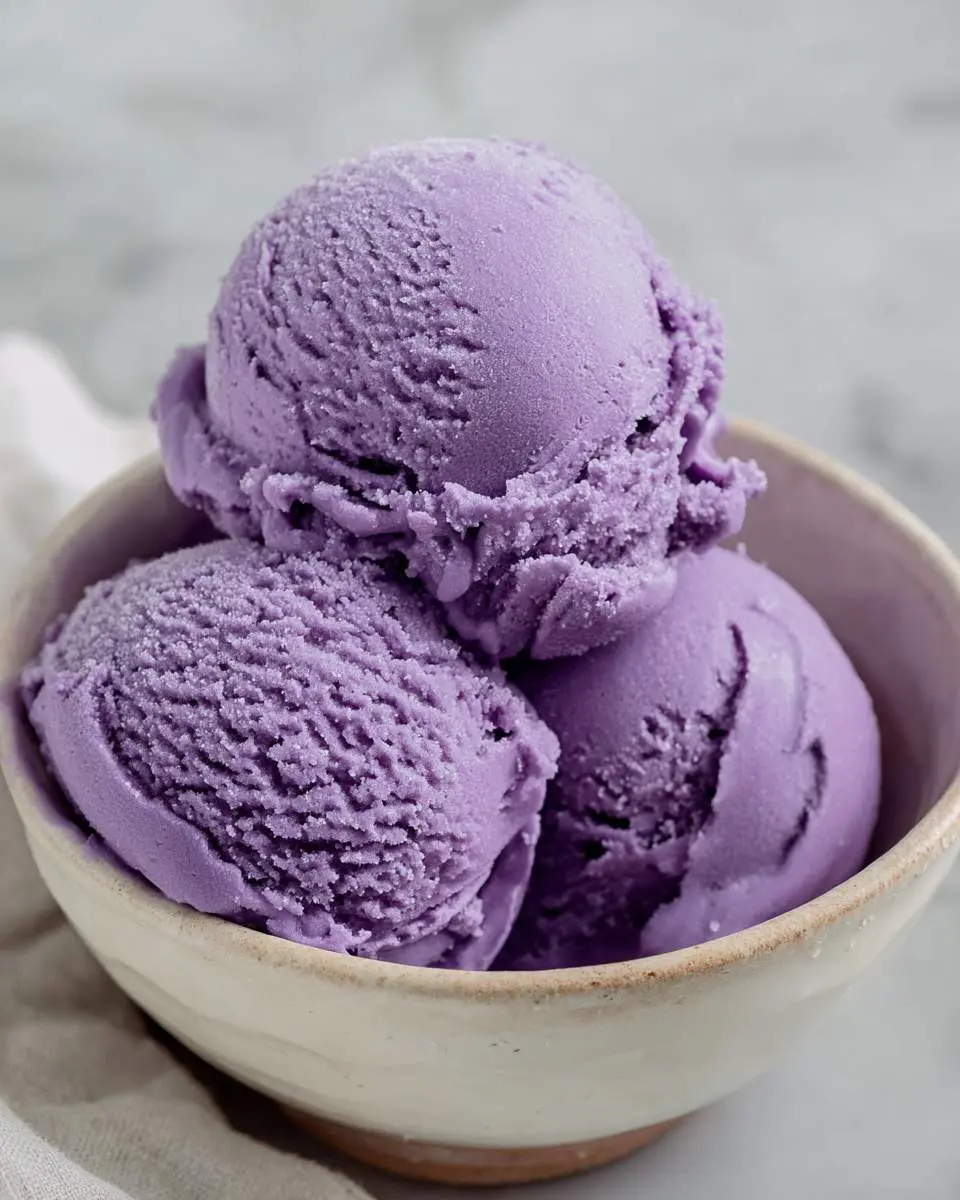Three scoops of creamy purple ube ice cream recipe served in a rustic ceramic bowl on a marble surface.