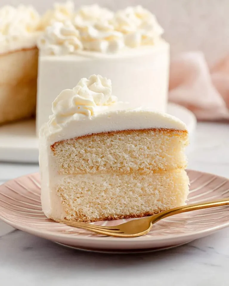 A slice of vanilla bean cake with white frosting on a pink plate with a gold fork.