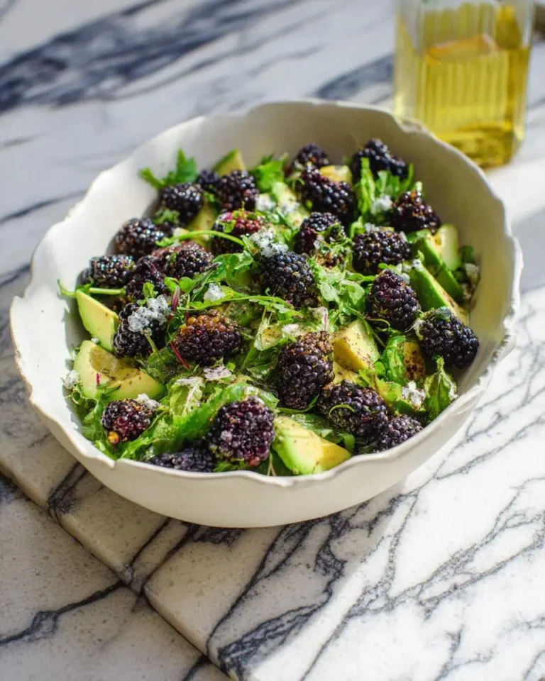 Fresh Blackberry Avocado Arugula Salad served in a white bowl with crumbled cheese and herbs on marble surface.