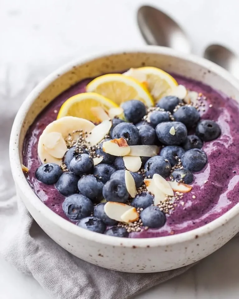 A creamy Blueberry Muffin Smoothie Bowl topped with fresh blueberries, lemon slices, banana, almond flakes, and chia seeds in a speckled bowl.