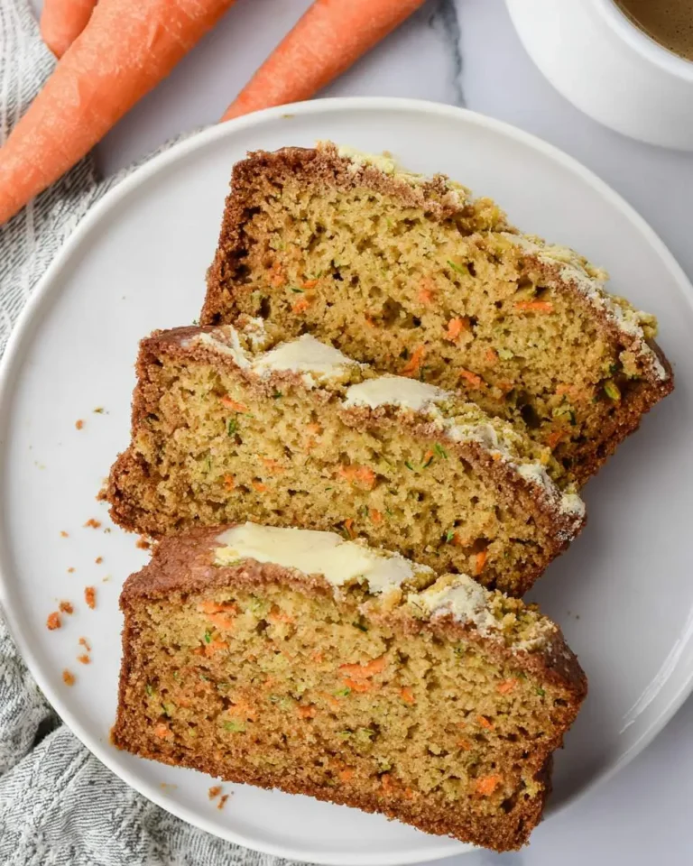 Three slices of Carrot Zucchini Bread with butter on a white plate, with fresh carrots in the background.