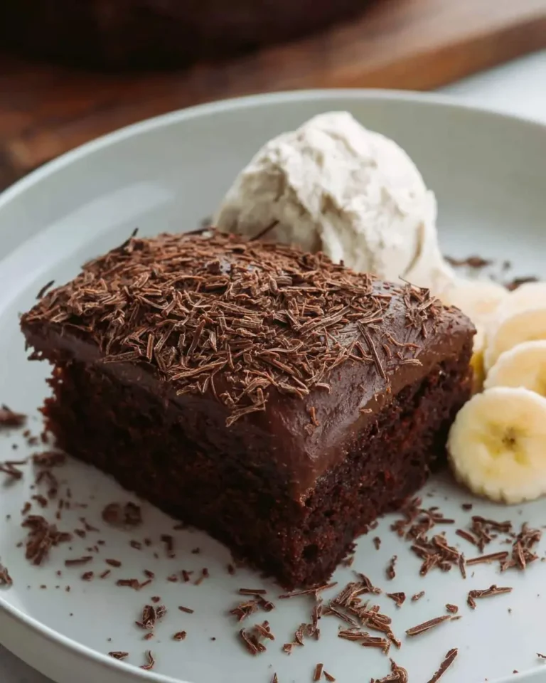 A slice of rich chocolate banana cake topped with chocolate shavings, served with whipped cream and fresh banana slices.