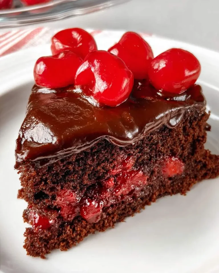 A slice of rich Chocolate Cherry Cake topped with glossy chocolate frosting and bright red cherries on a white plate.