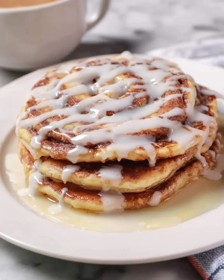 A stack of Cinnamon Roll Pancakes drizzled with white icing on a white plate, with a cup of coffee in the background.