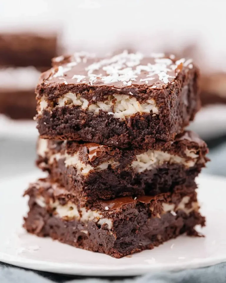 Three coconut brownies stacked on a white plate, topped with chocolate glaze and shredded coconut.