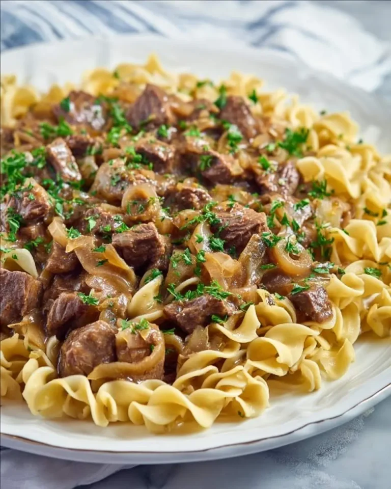 A plate of crockpot french onion beef and noodles topped with fresh parsley, featuring tender beef chunks and caramelized onions over egg noodles.