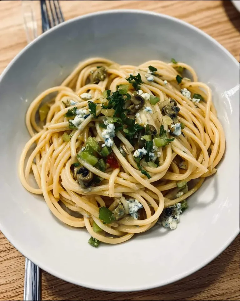 A bowl of Dirty Martini Spaghetti topped with blue cheese crumbles, chopped herbs, and olives on a wooden table.