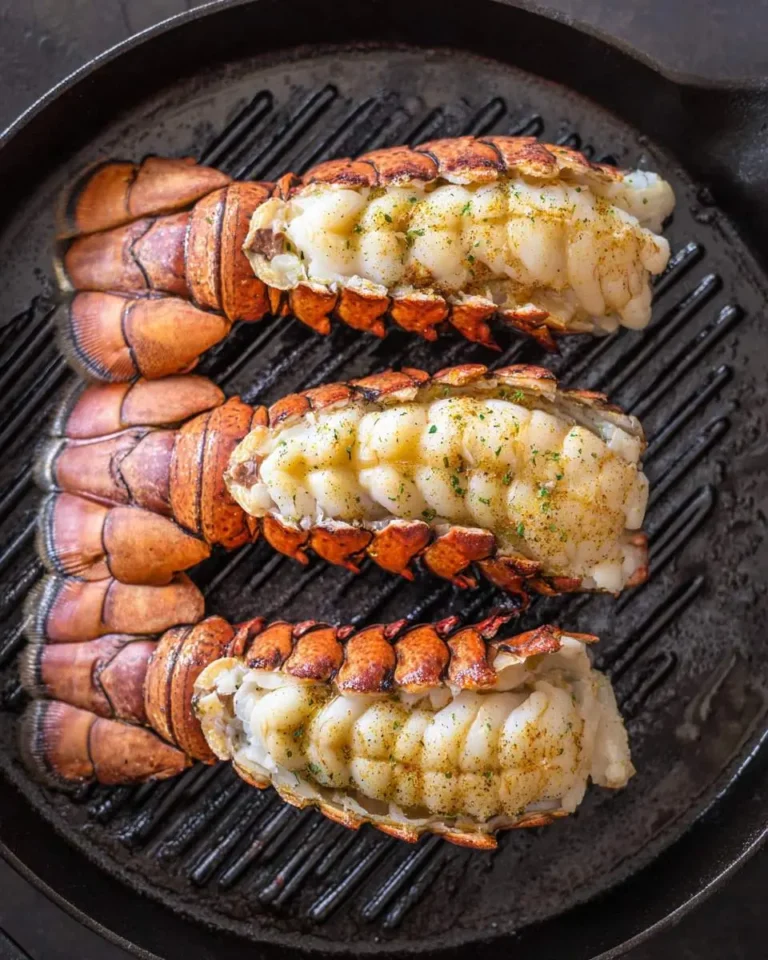 Three grilled lobster tails seasoned with herbs cooking on a cast iron grill pan.