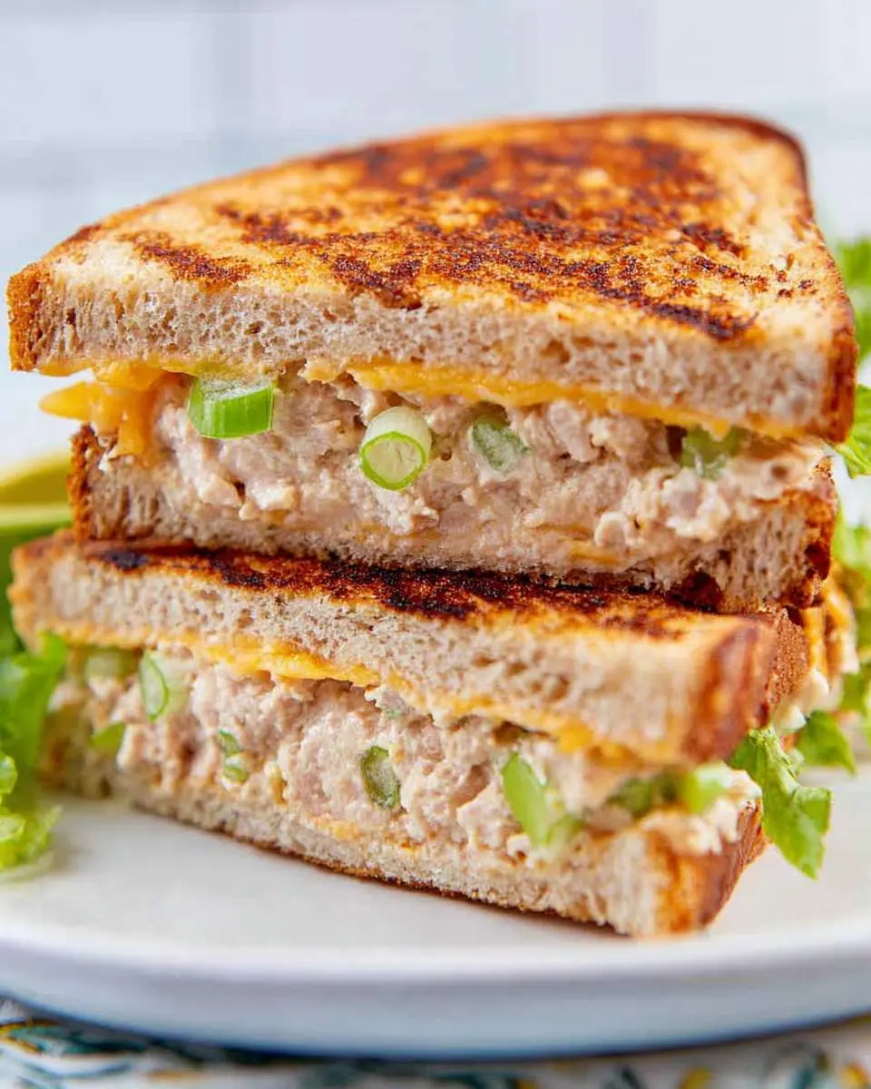 Two halves of a grilled tuna sandwich stacked on a white plate, showing melted cheese and green onions inside.