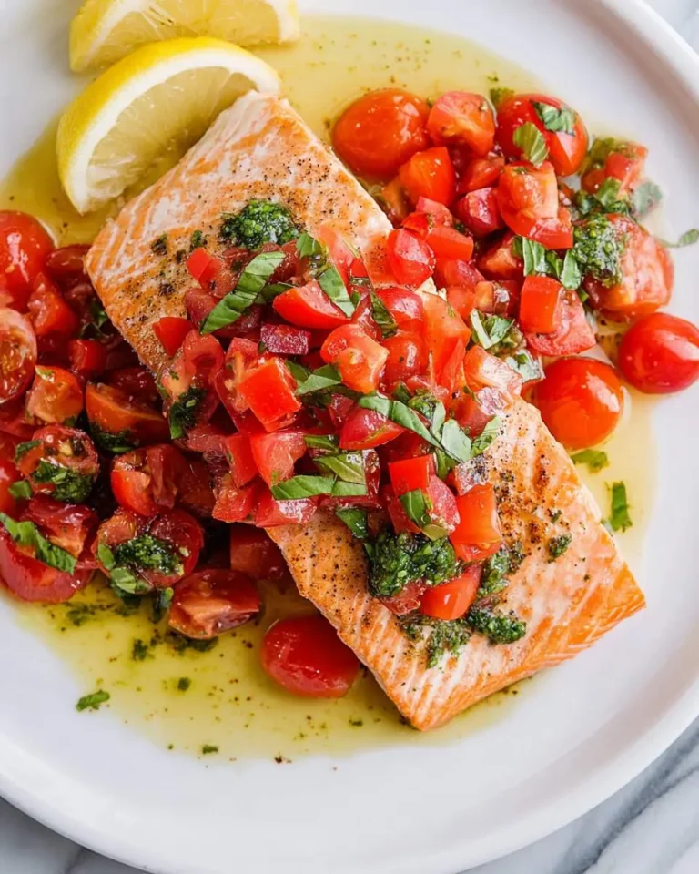 Pan-seared salmon fillets topped with fresh basil sauce and vibrant tomato salad, served with lemon wedges on a white plate.