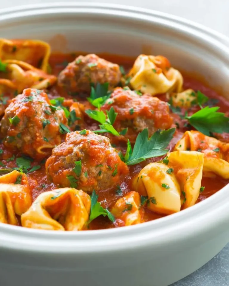 A white slow cooker filled with Slow Cooker Tortellini Meatballs in rich tomato sauce, garnished with fresh parsley.