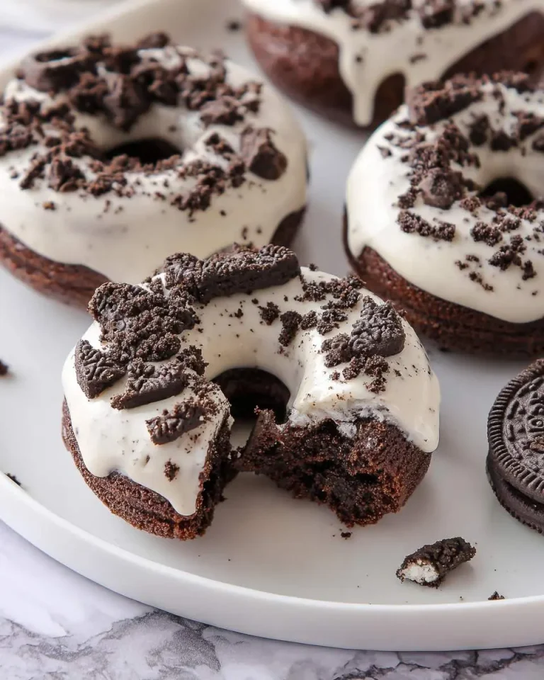 Baked Oreo Donuts with white glaze and cookie crumbles on a white plate, one with a bite taken out.