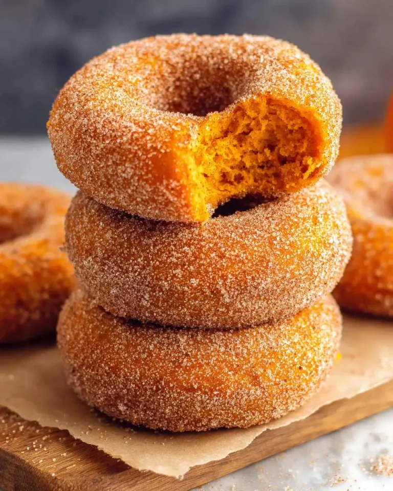 Stack of three baked pumpkin donuts coated in cinnamon sugar, with the top donut showing a bite taken out.