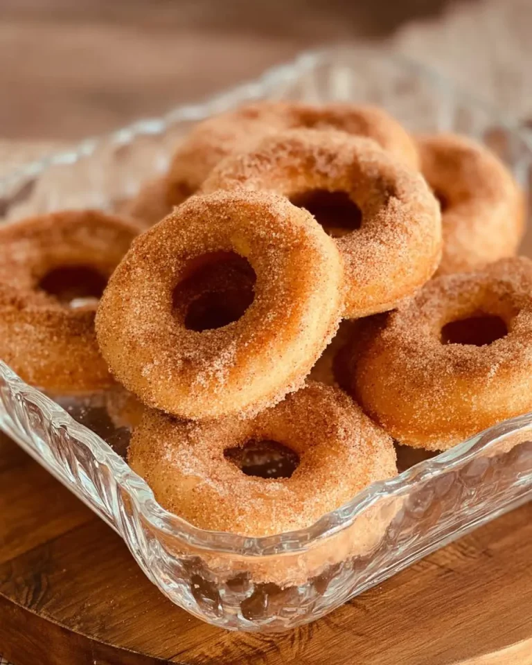 A glass dish filled with Churro Baked Donuts coated in cinnamon sugar, stacked on a wooden surface.