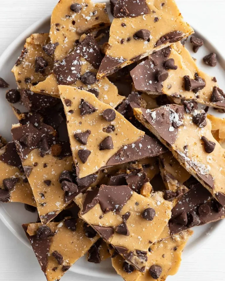 A white plate piled high with irregular pieces of Cookie Dough Bark topped with chocolate chunks and flaky sea salt.