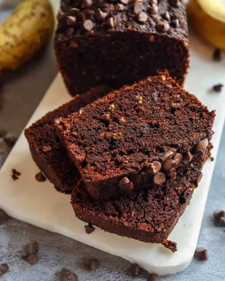 Sliced Double Chocolate Banana Bread topped with chocolate chips on a white cutting board with bananas in background.