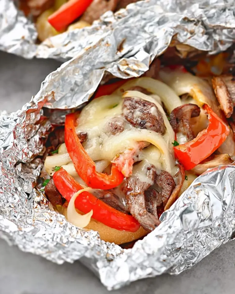 A foil pack with Philly cheese steak open to reveal melted cheese, sliced beef, peppers, and onions on a toasted bun.