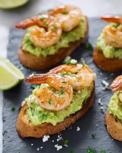 Garlic Shrimp and Avocado Crostini on toasted bread with lime wedges and fresh herbs on a slate board.