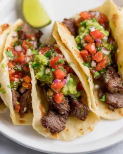 Three Mexican Steak Tacos on a white plate topped with fresh guacamole, pico de gallo, and a lime wedge in the background.