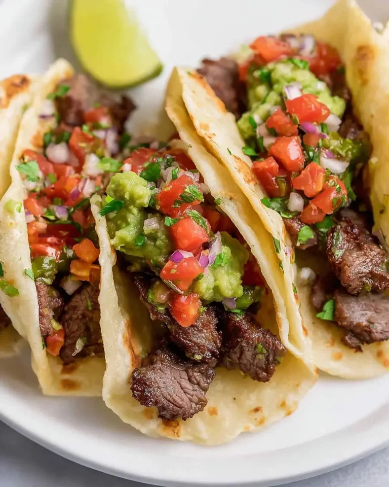Three Mexican Steak Tacos on a white plate topped with fresh guacamole, pico de gallo, and a lime wedge in the background.