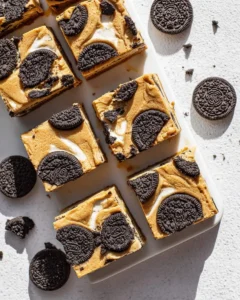 Squares of Peanut Butter Oreo Bars arranged on a white surface with whole and crushed Oreo cookies scattered around.