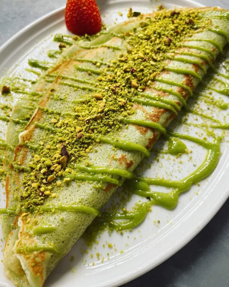 Two pistachio crepes drizzled with green sauce and topped with crushed pistachios, served with a strawberry.