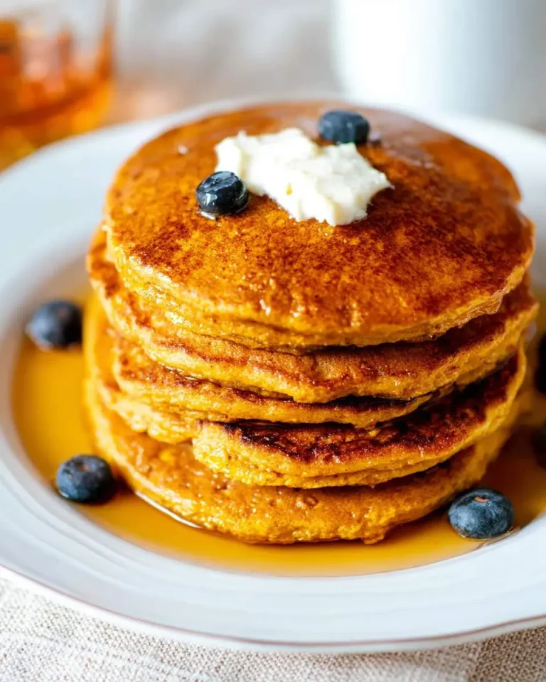 Stack of golden pumpkin pancakes topped with butter, blueberries, and maple syrup on a white plate.