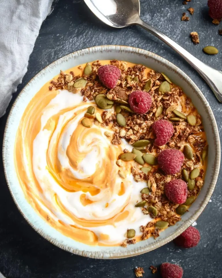 Pumpkin Pie Yogurt Bowl topped with raspberries, granola, and pumpkin seeds on a dark surface.