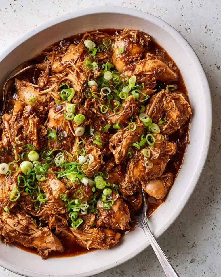 Slow Cooker Hoisin Chicken shredded in a white bowl, topped with sliced green onions and rich brown sauce.