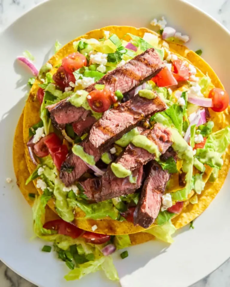 Steak Tostada Salad with crispy tortillas, grilled steak strips, fresh vegetables, and avocado dressing on a white plate.