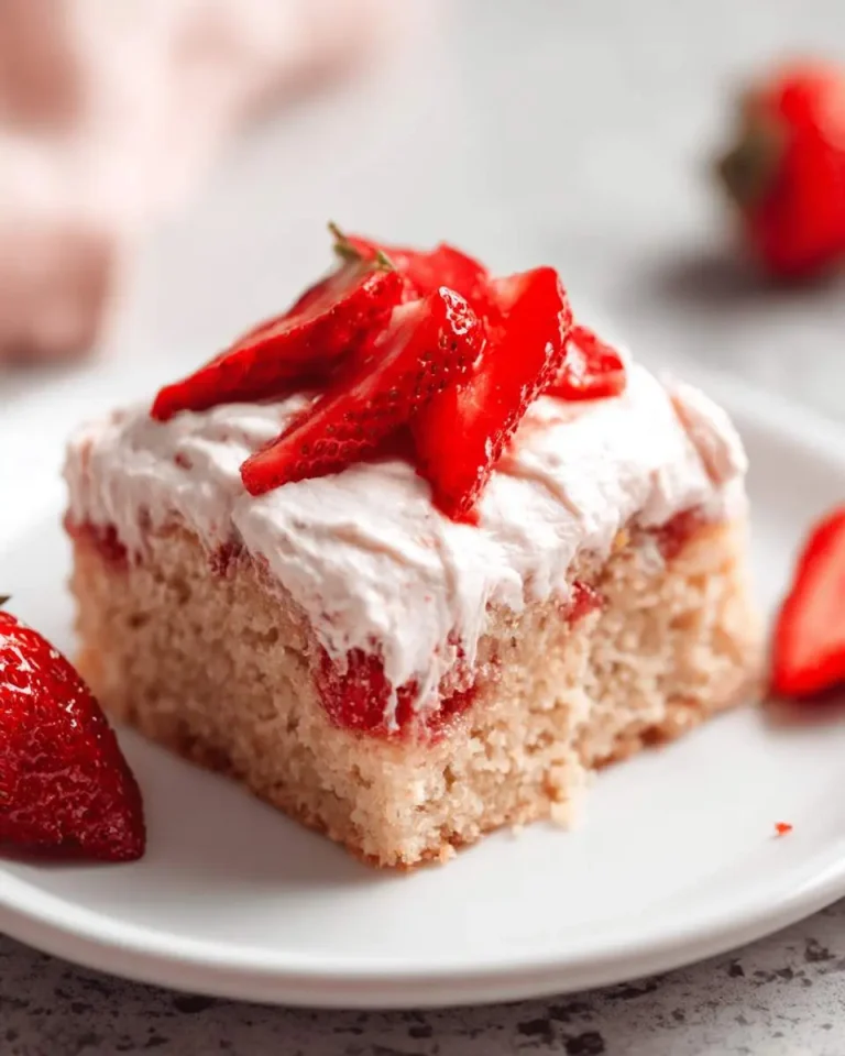 A slice of Strawberry Sheet Cake topped with creamy frosting and fresh strawberry slices on a white plate.