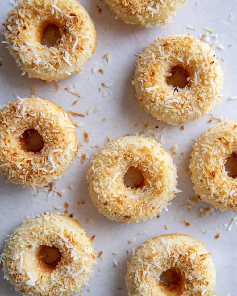 Homemade Toasted Coconut Donuts arranged on parchment paper, topped with golden brown shredded coconut flakes.