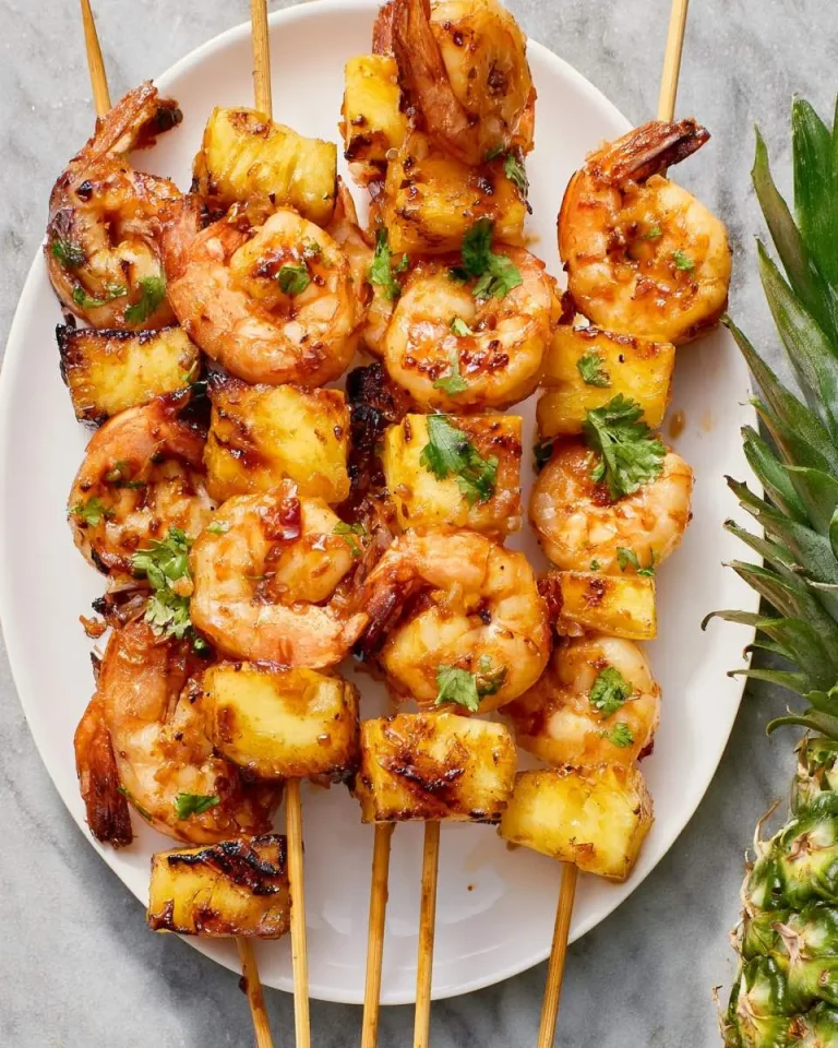 Grilled Asian Shrimp Pineapple Skewers on a white plate, garnished with fresh cilantro and a whole pineapple beside it.