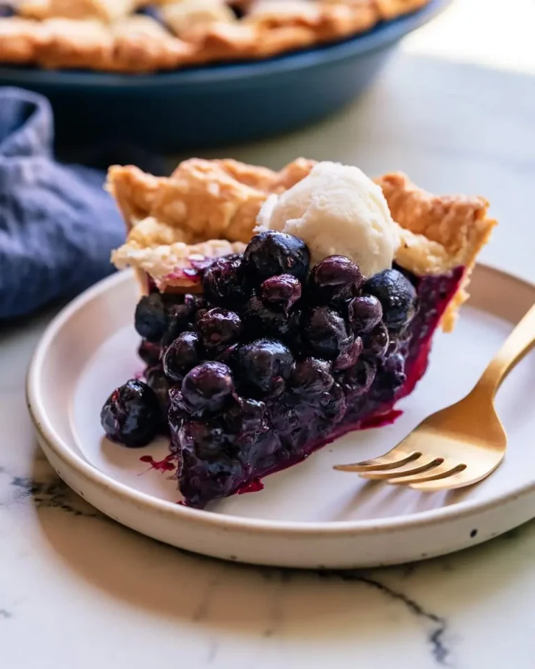 A slice of homemade Blueberry Pie topped with vanilla ice cream on a white plate with a gold fork.