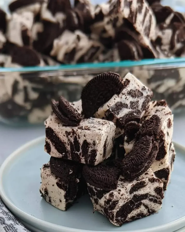 Stack of creamy Oreo Fudge squares topped with cookie pieces on a blue plate with more fudge in background.